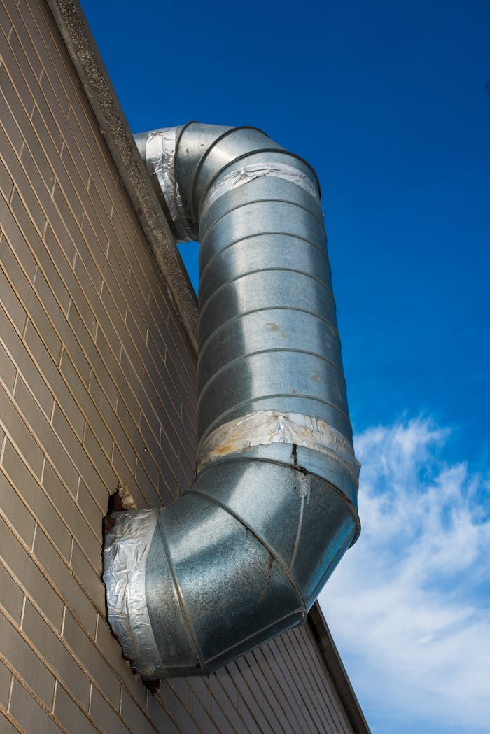 Home A large industrial air duct attached to a brick wall under a bright blue sky.